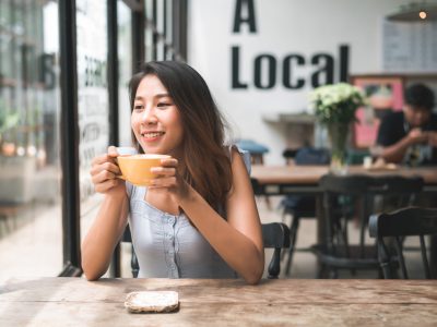 Cheerful asian young woman drinking warm coffee or tea enjoying it while sitting in cafe. Attractive happy asian woman holding a cup of coffee.