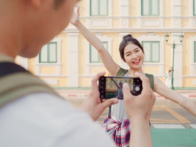 Traveler Asian couple travel in Bangkok, Thailand, sweet couple using mobile phone taking photo each other near street while spending holiday trip in sunset. Lifestyle couple travel in city concept.